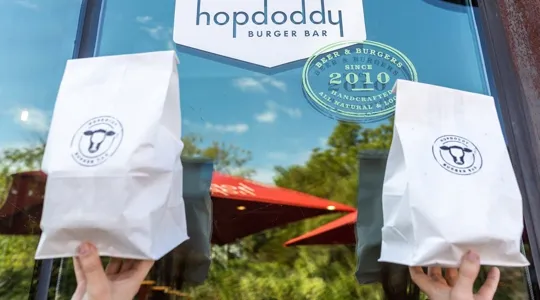 Hands holding up two Hopdoddy to-go bags in front of Hopdoddy sign