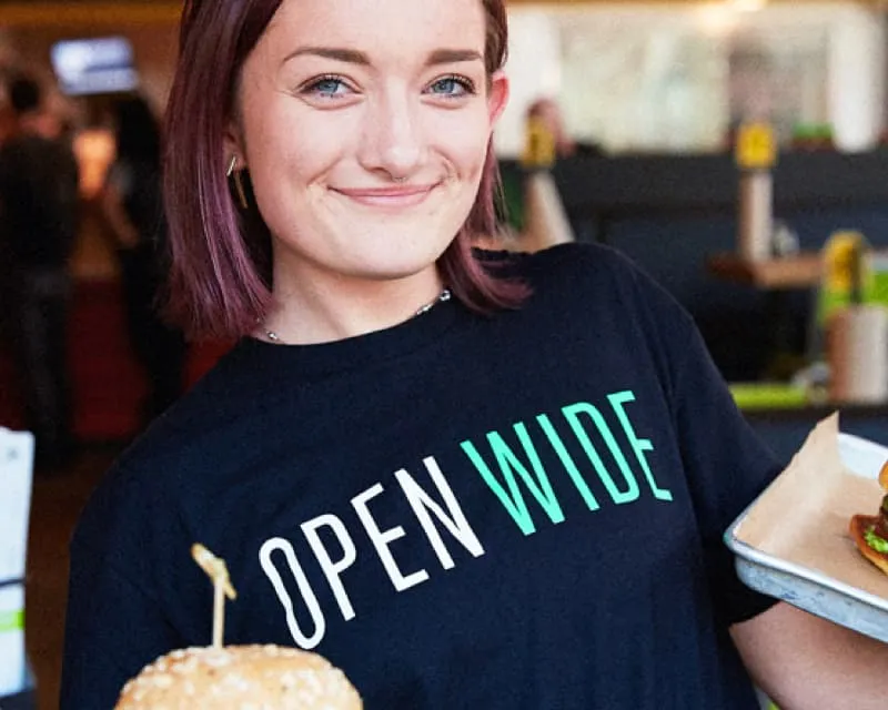 Employee smiling and holding two big burgers while wearing an Open Wide Hopdoddy t-shirt